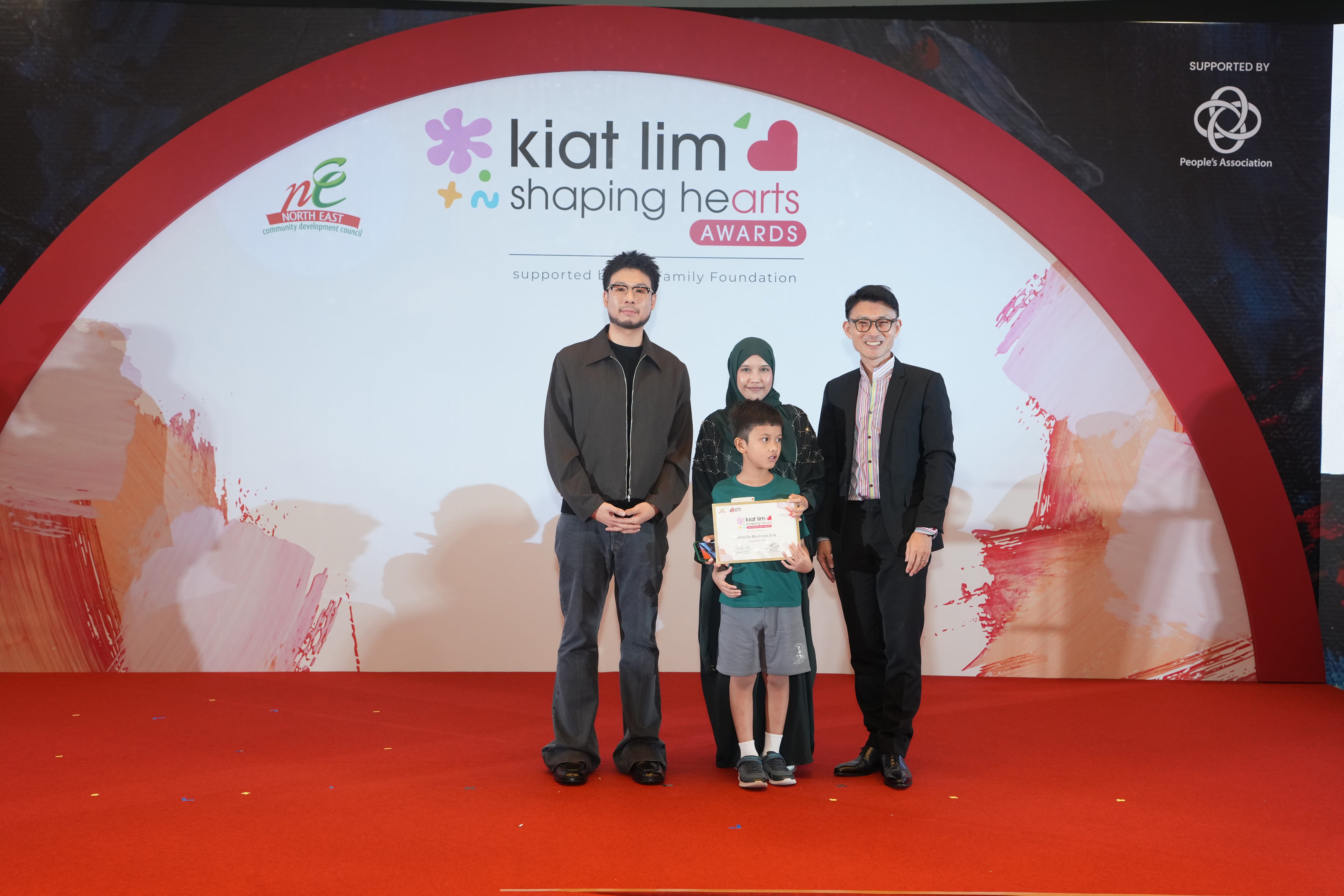 Award recipient standing on stage with the presenter, holding their certificate and posing for a group photo during the award ceremony, with a backdrop reading ‘Kiat Lim-Shaping Hearts Awards'.